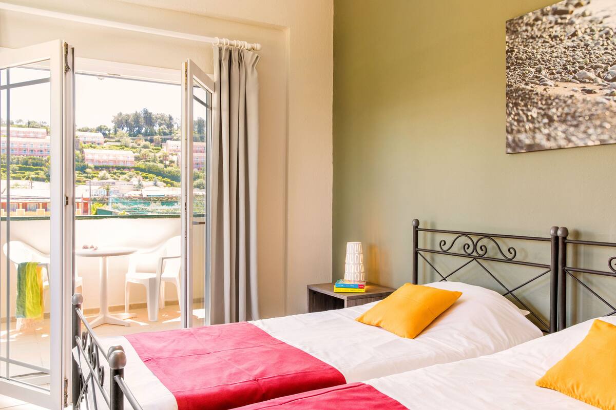 A hotel room with a balcony, a bed with white and red bedding, and a view of buildings and greenery outside.