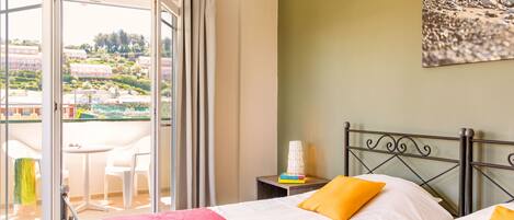 A hotel room with a balcony, a bed with white and red bedding, and a view of buildings and greenery outside.