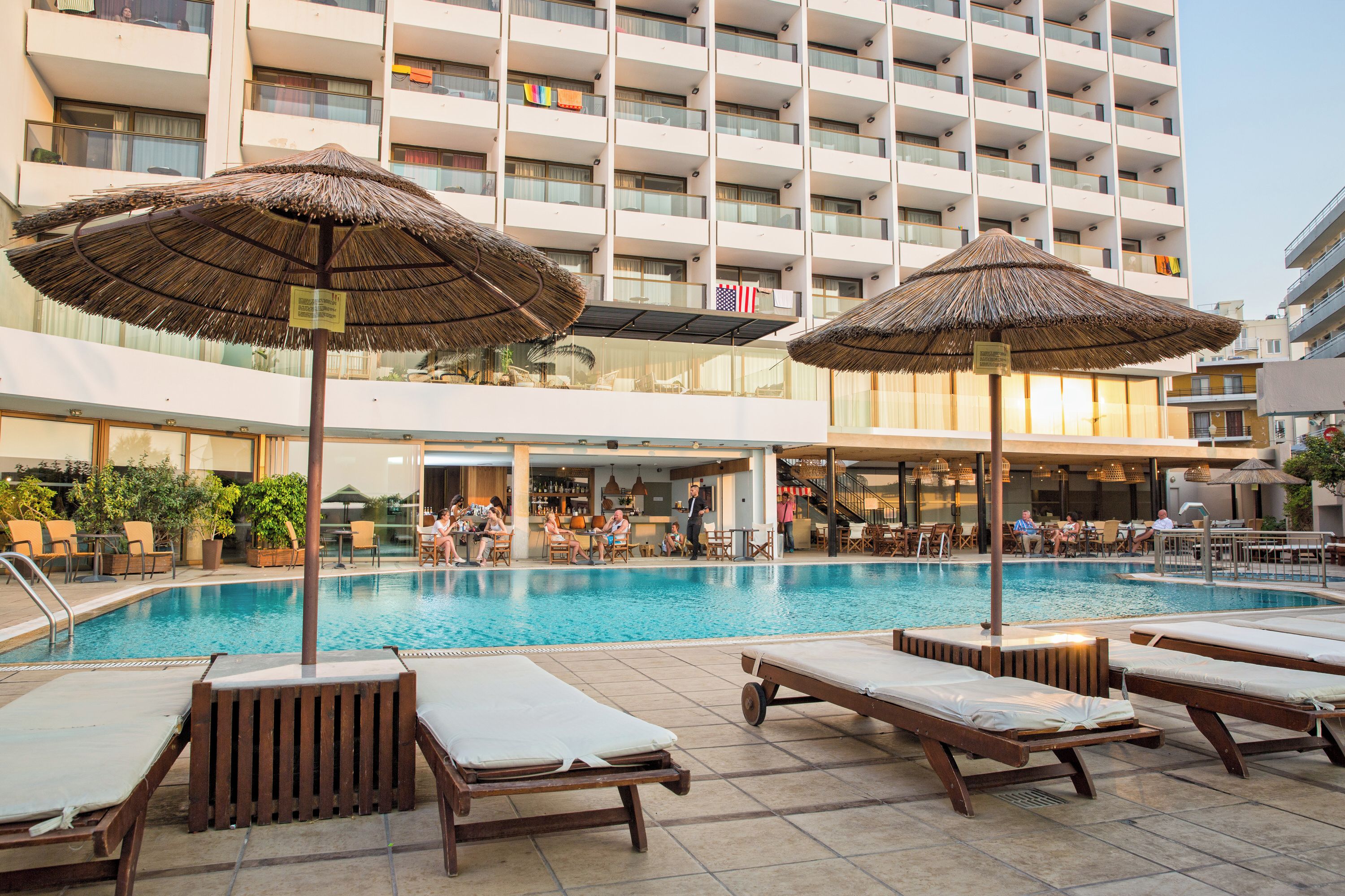 A hotel pool area with lounge chairs and umbrellas, a bar, and a view of the building's exterior.