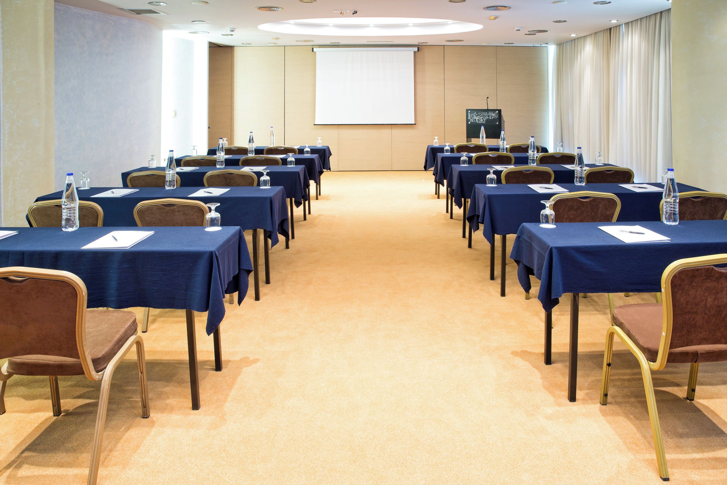 Konferenssihuone, jossa on riveissä pöytiä ja tuoleja, projektorikangas ja matolla peitetty lattia.