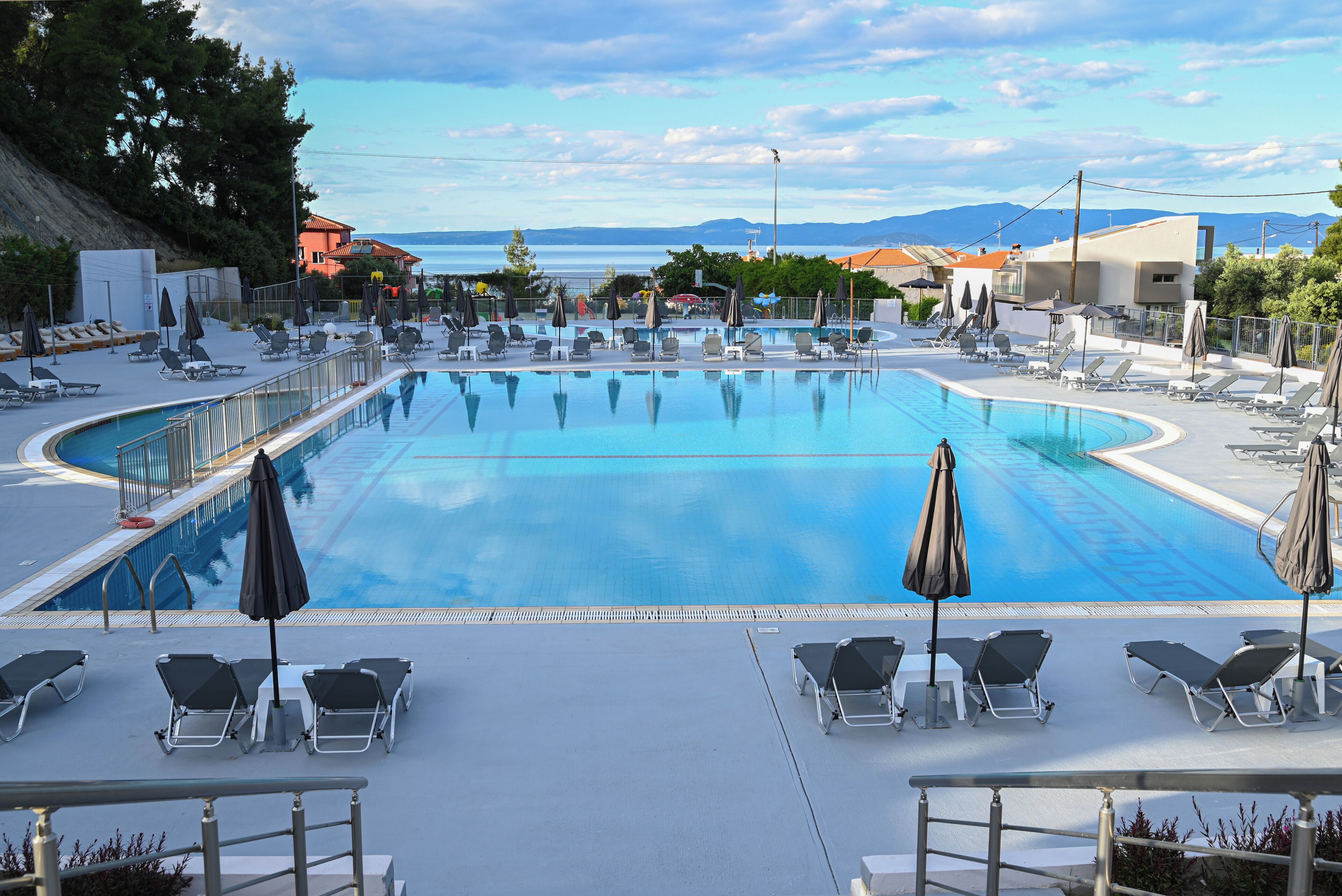 A large swimming pool with lounge chairs and umbrellas, surrounded by buildings and a view of the sea.