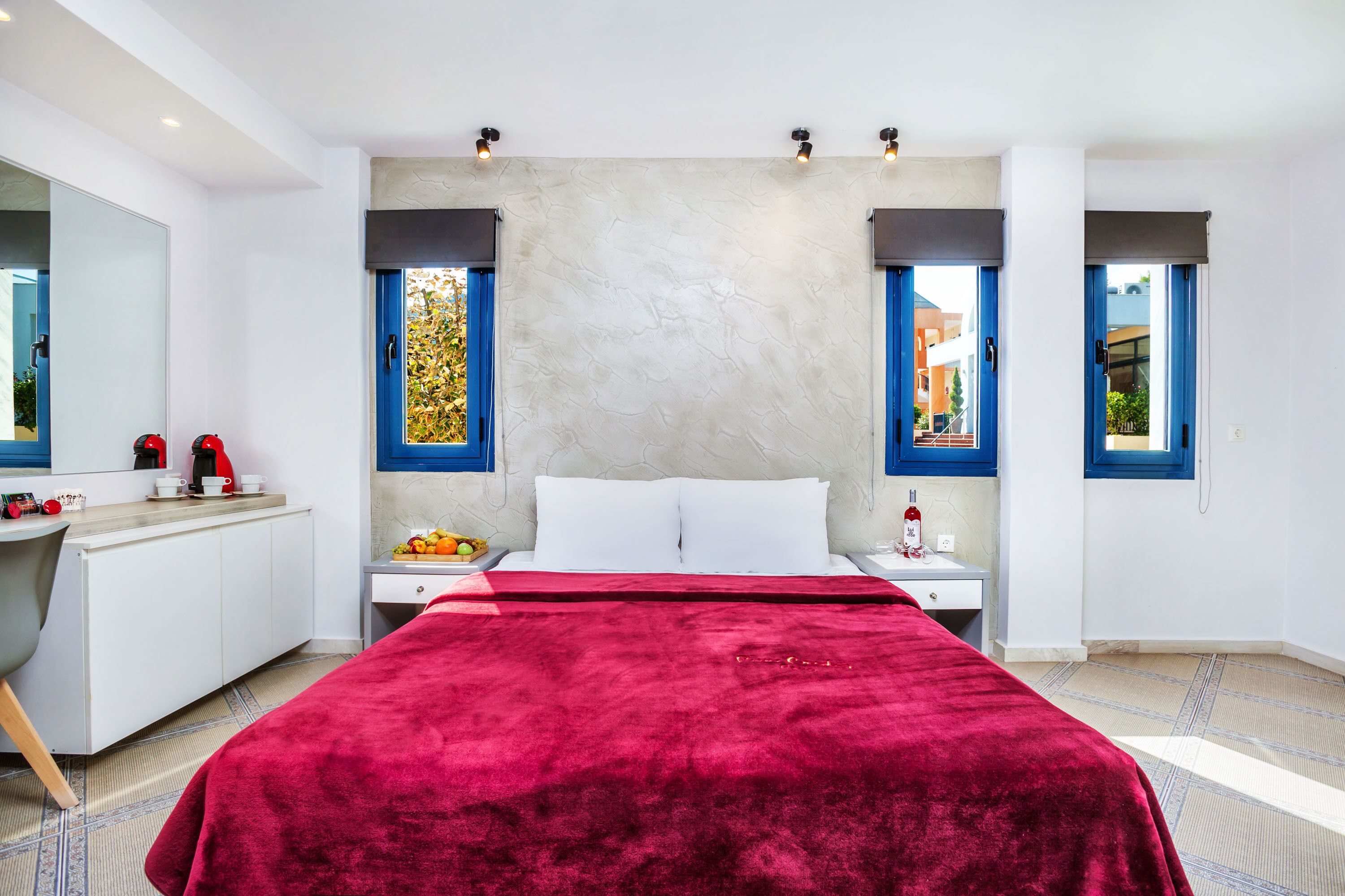 A modern hotel room with a large bed, a bedside table with a bottle, a fruit bowl, and a red blanket.