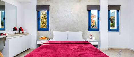 A modern hotel room with a large bed, a bedside table with a bottle, a fruit bowl, and a red blanket.