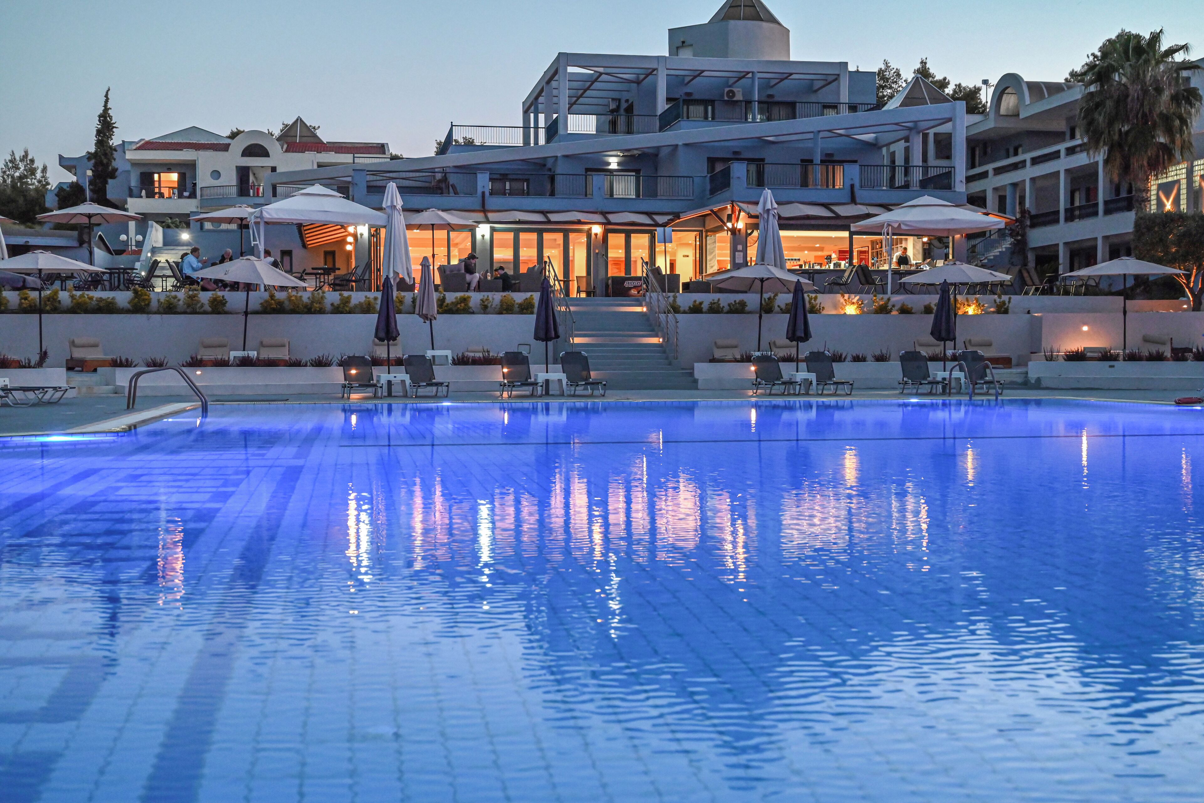 A swimming pool with lounge chairs and umbrellas, a multi-story building with balconies, and a well-lit entrance.