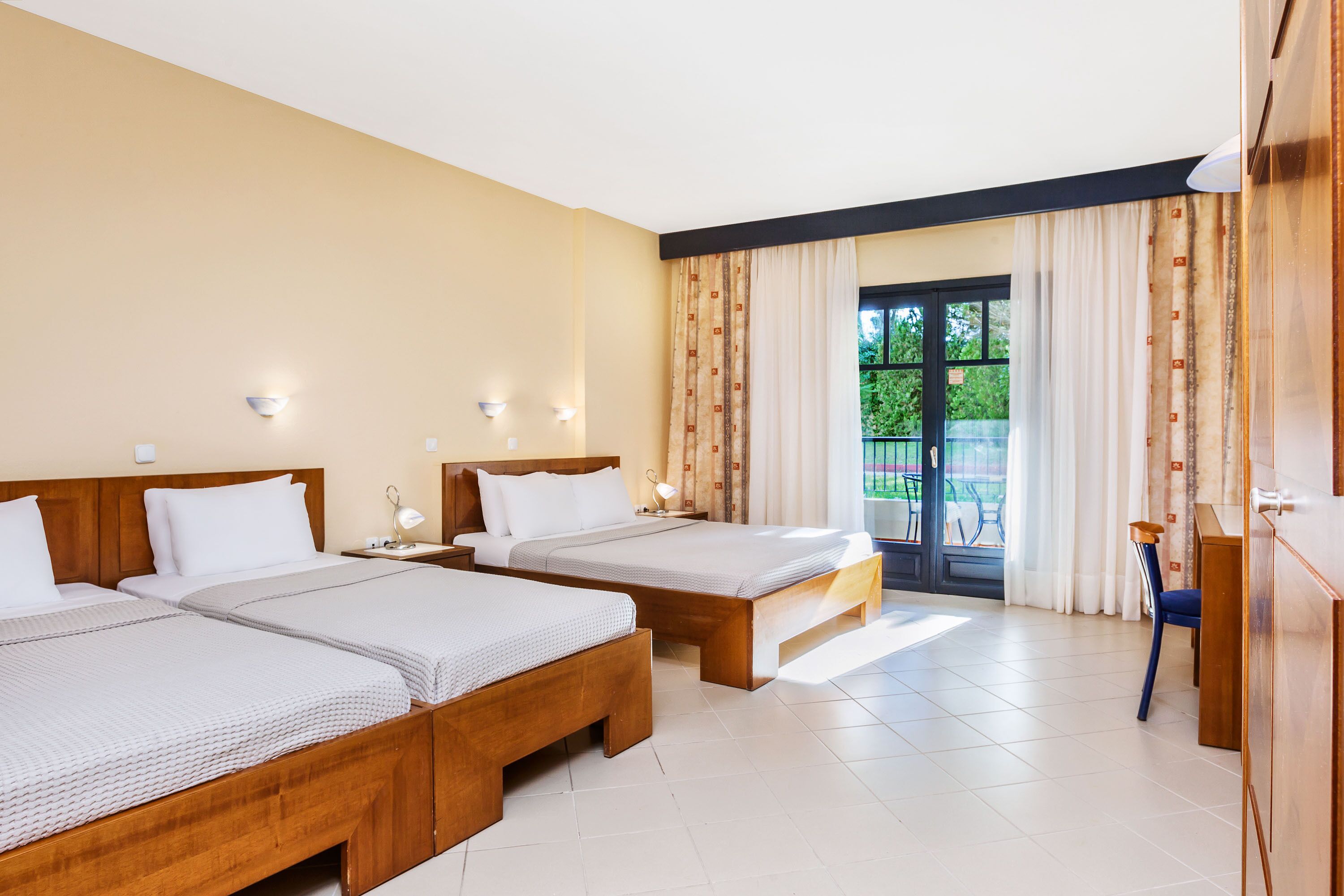 A hotel room with two wooden beds, a nightstand, a desk, and a view of the outdoors through a large window.