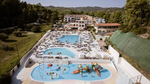 A large outdoor swimming pool with multiple lanes, surrounded by lounge chairs and umbrellas, and a playground area with slides and water features.