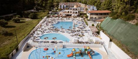 A large outdoor swimming pool with multiple lanes, surrounded by lounge chairs and umbrellas, and a playground area with slides and water features.