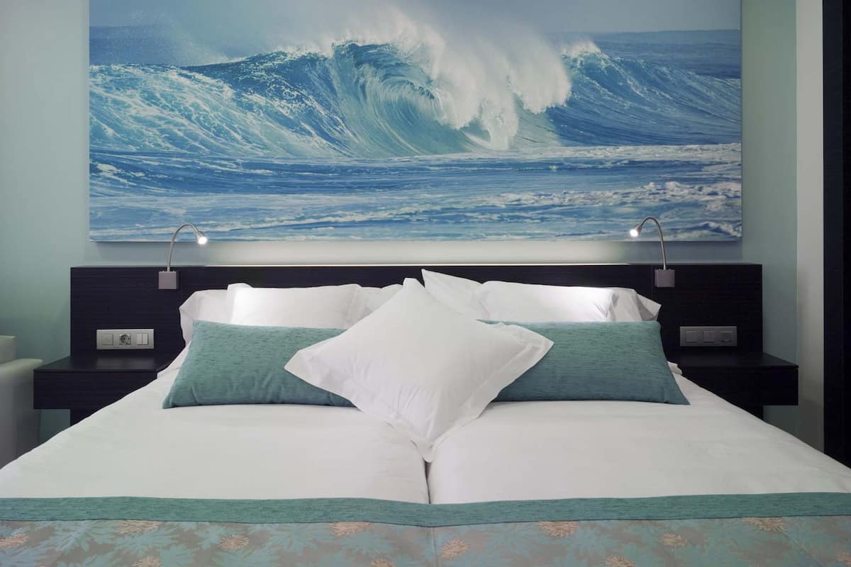 A bedroom with a large bed, two bedside tables, and a wall-mounted painting of a wave.