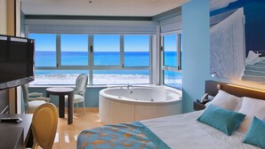A hotel room with a large bed, a bathtub, a TV, a desk, and a chair. There are large windows with a view of the beach and ocean. - Villa del Mar Hotel (Benidorm)