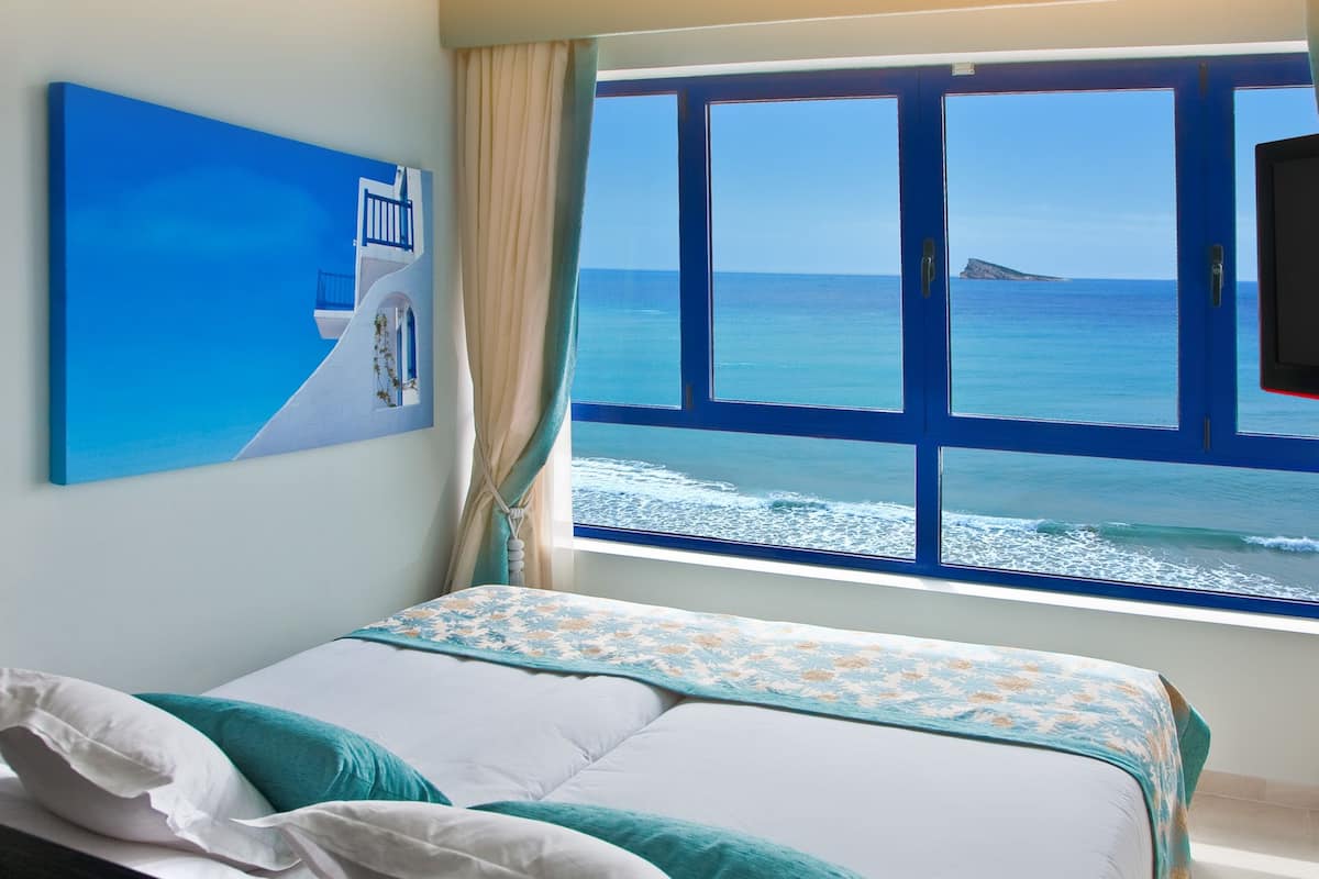A bedroom with a bed, a window with a view of the sea, and a painting on the wall.