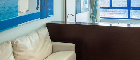 A modern room with a beige sofa, a dark wooden desk, and a large window offering a view of the sea.