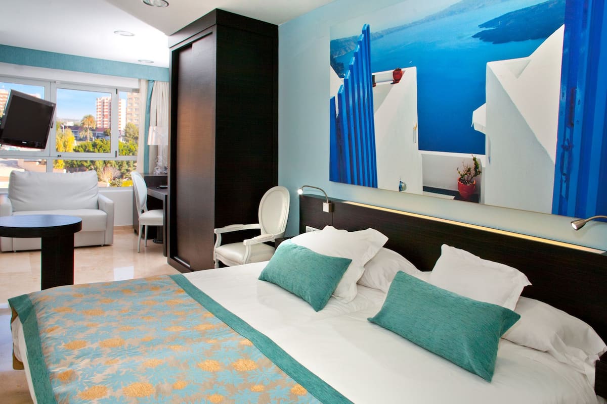 A hotel room with a large bed, two chairs, a desk, and a view of the city through large windows.