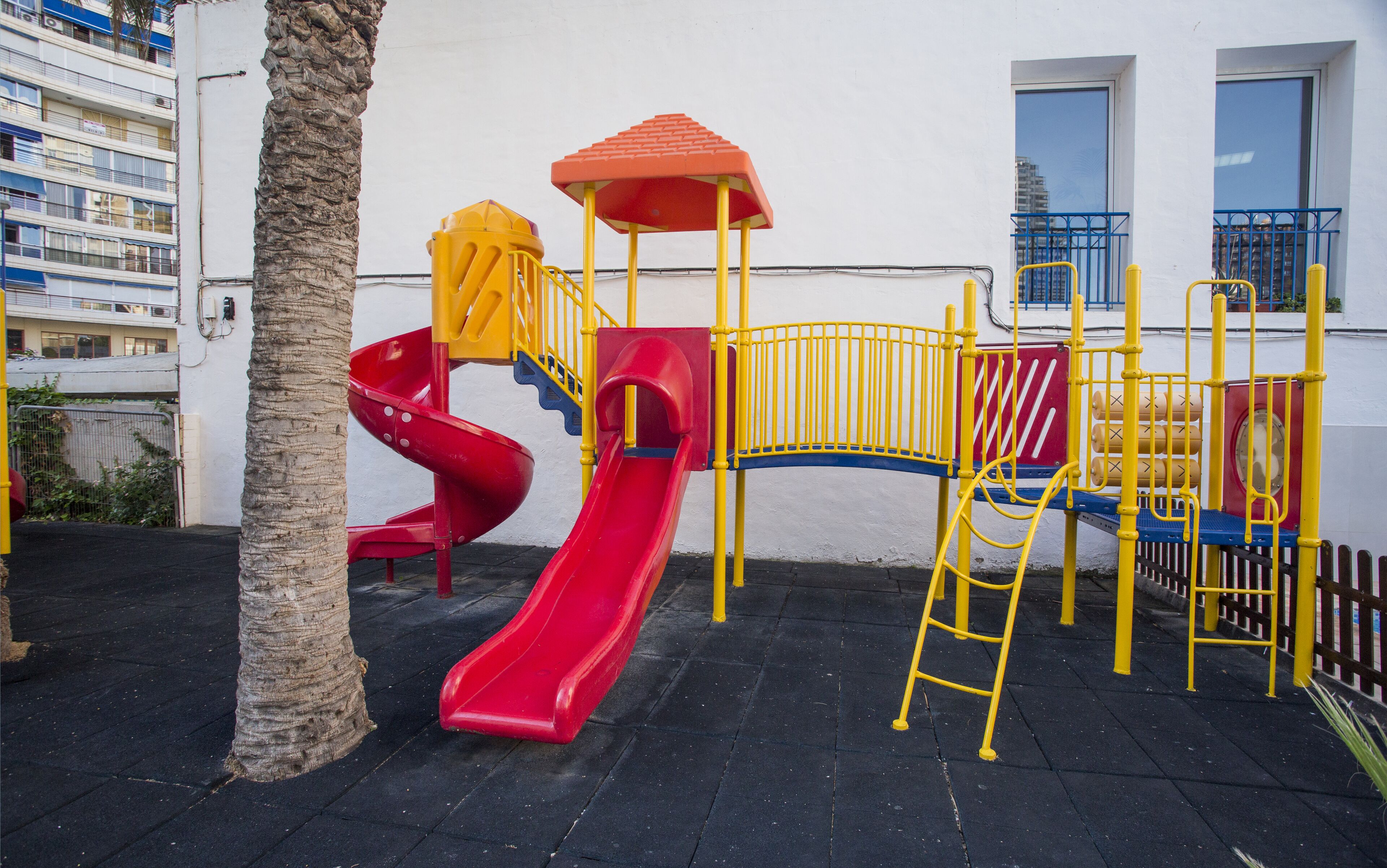 Een speeltuin met een rode glijbaan, gele klimconstructies en een overkapping met een rood dak.