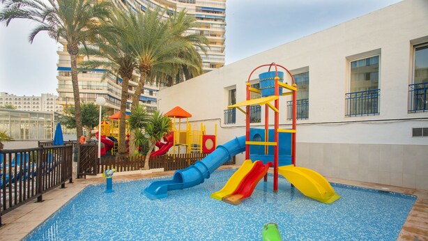 A children's play area with slides and a climbing structure, surrounded by a pool and palm trees.