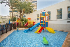 A children's play area with slides and a climbing structure, surrounded by a pool and palm trees.