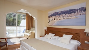 A hotel room with a large bed, a seating area with a chair, a window with curtains, and a painting of a cityscape.