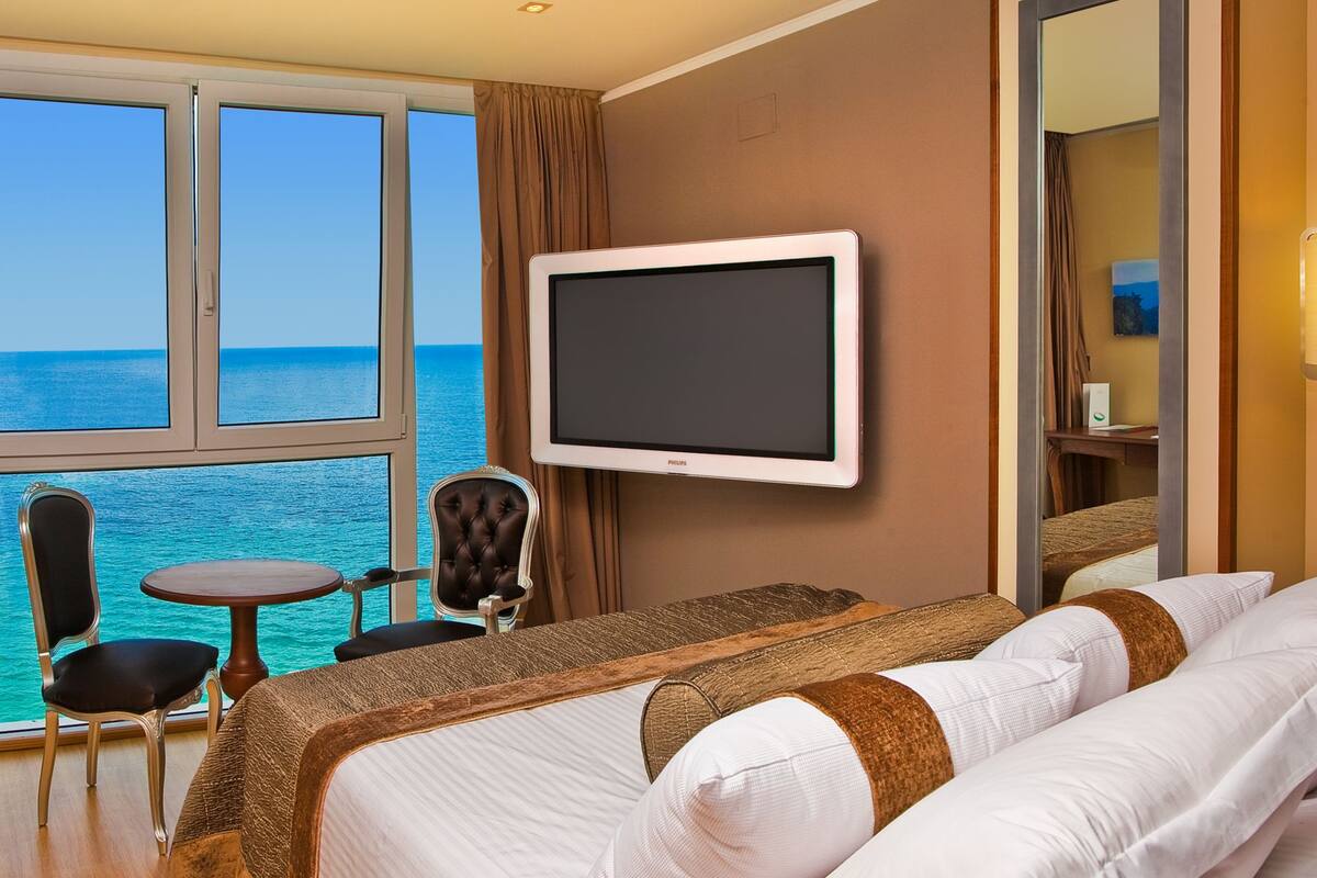 Ein Hotelzimmer mit großem Fenster und Meerblick, einem Bett mit weißer Bettwäsche, einem Fernseher und einem kleinen Tisch mit zwei Stühlen.