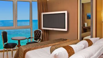 Habitación de hotel con un ventanal amplio que ofrece vistas al mar, cama con ropa blanca, televisor y una pequeña mesa con dos sillas.