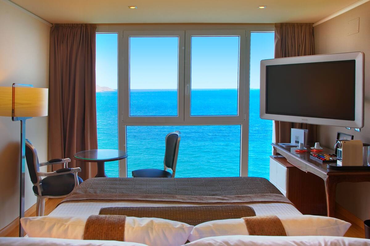 Ein Hotelzimmer mit einem großen Bett, einem Schreibtisch, einem Flachbildfernseher, einem Sessel und Meerblick durch große Fenster.