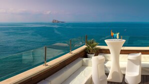 Une terrasse dotée de meubles blancs, d’une plante en pot et offrant une vue sur l’océan et une île lointaine.