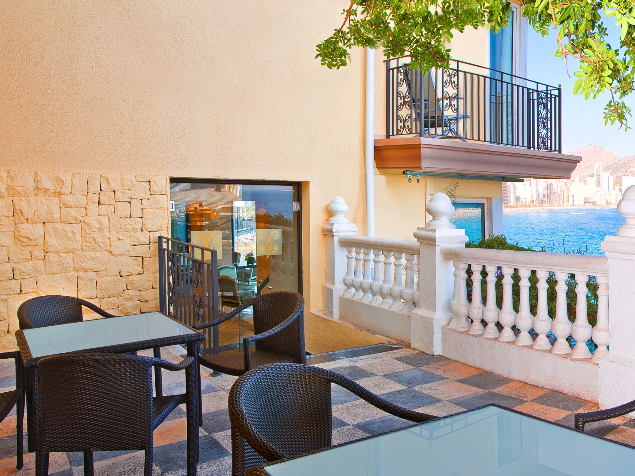 An outdoor seating area with wicker chairs and a glass table, overlooking a body of water and a cityscape.
