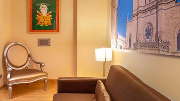 Habitación de hotel con un sofá de cuero marrón, una silla de estructura metálica, una lámpara y un cuadro de un edificio en la pared.