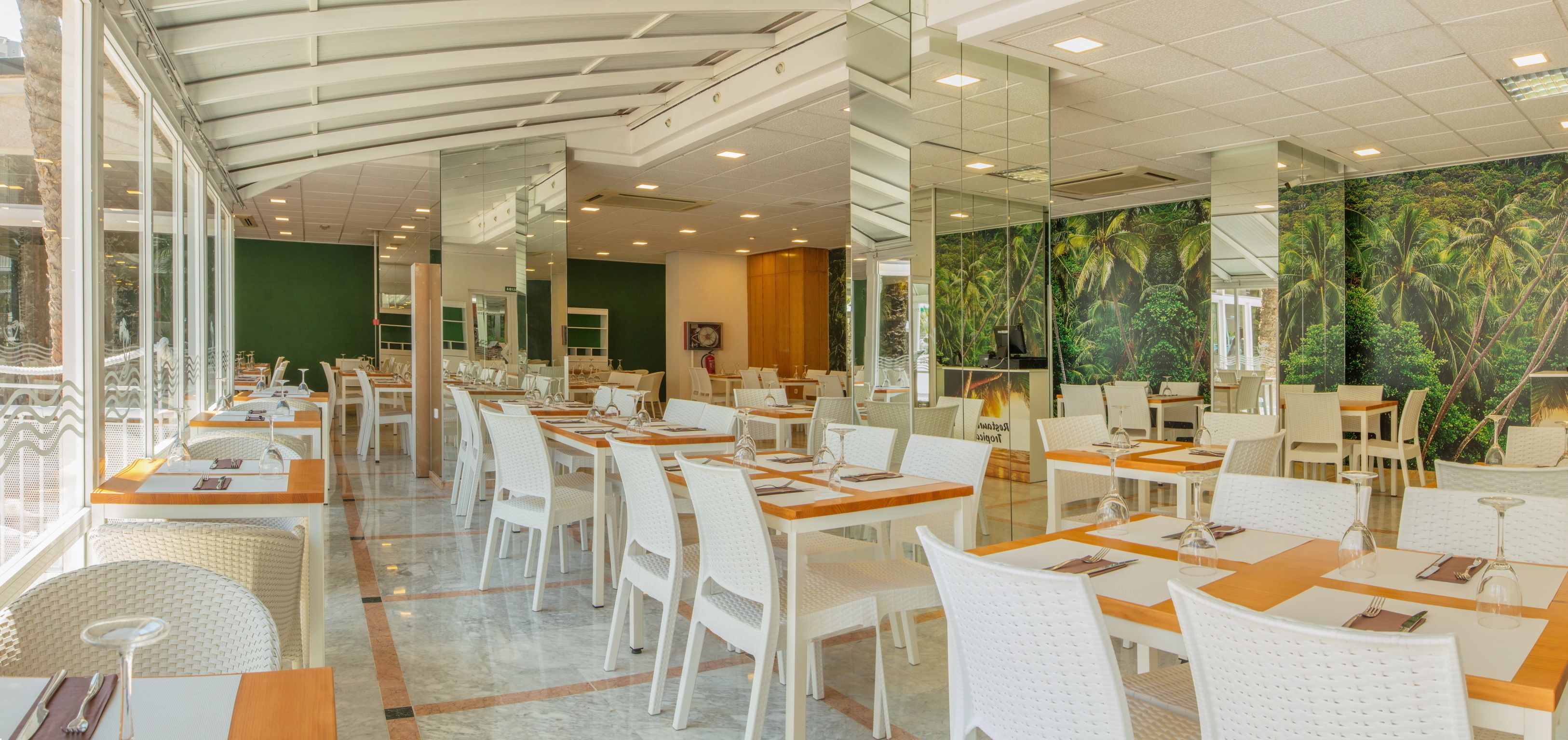 Un restaurante espacioso con sillas y mesas blancas, ventanas amplias y un mural tropical en la pared.
