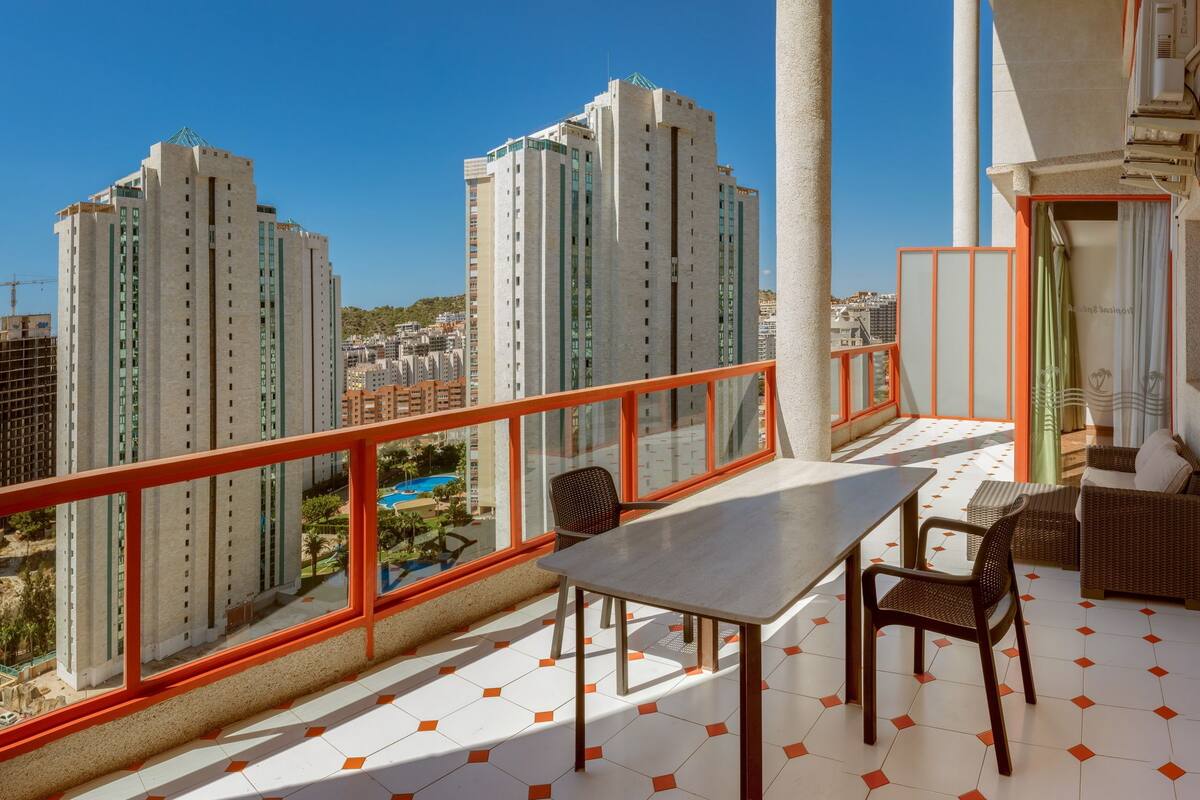 Un balcón con mesa y sillas, vista a edificios altos y una piscina al fondo.