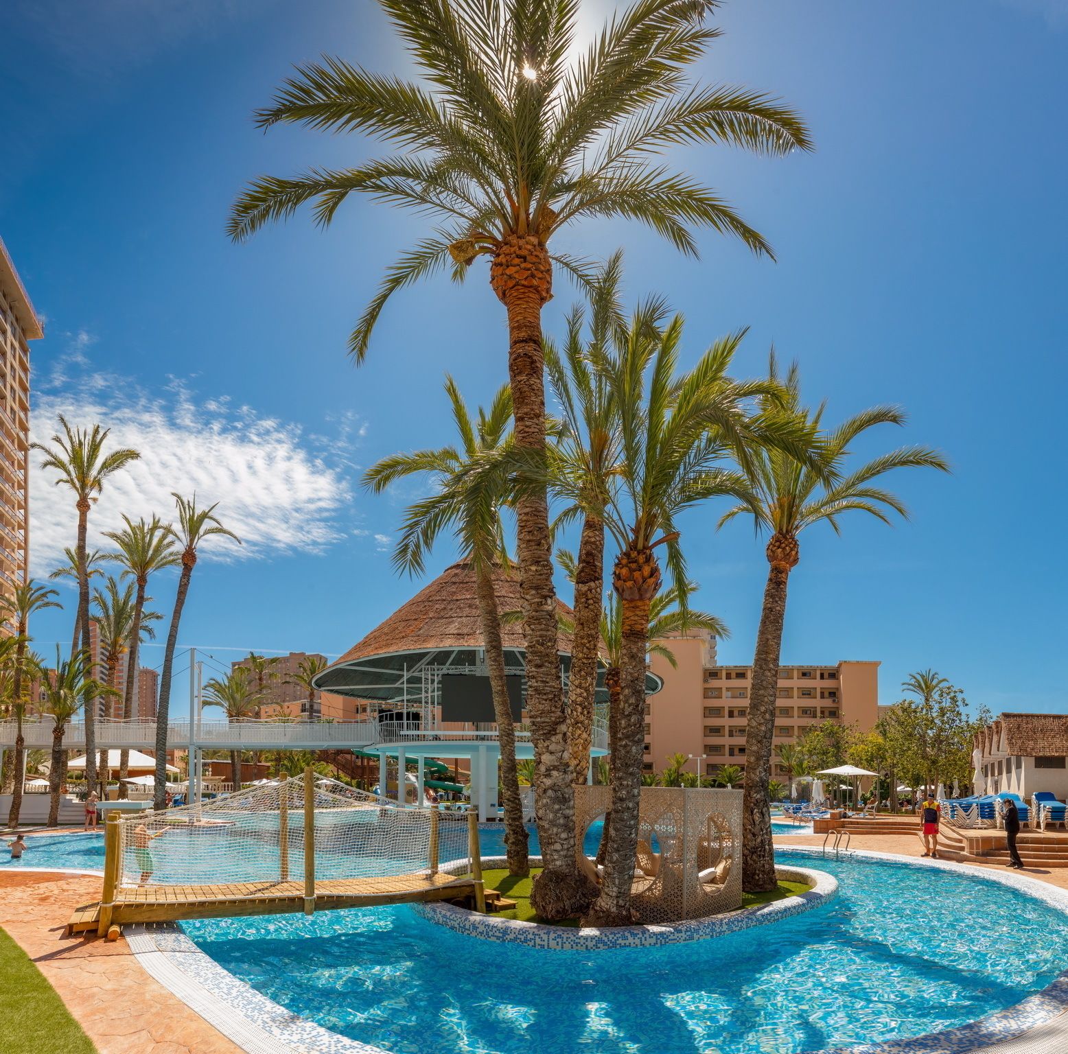 Un espace piscine agrémenté de palmiers, un coin salon abrité et un bâtiment en arrière-plan.
