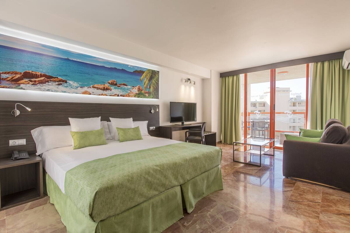 Habitación de hotel con cama, escritorio, televisor y balcón con cortinas, además de un cuadro con una playa.