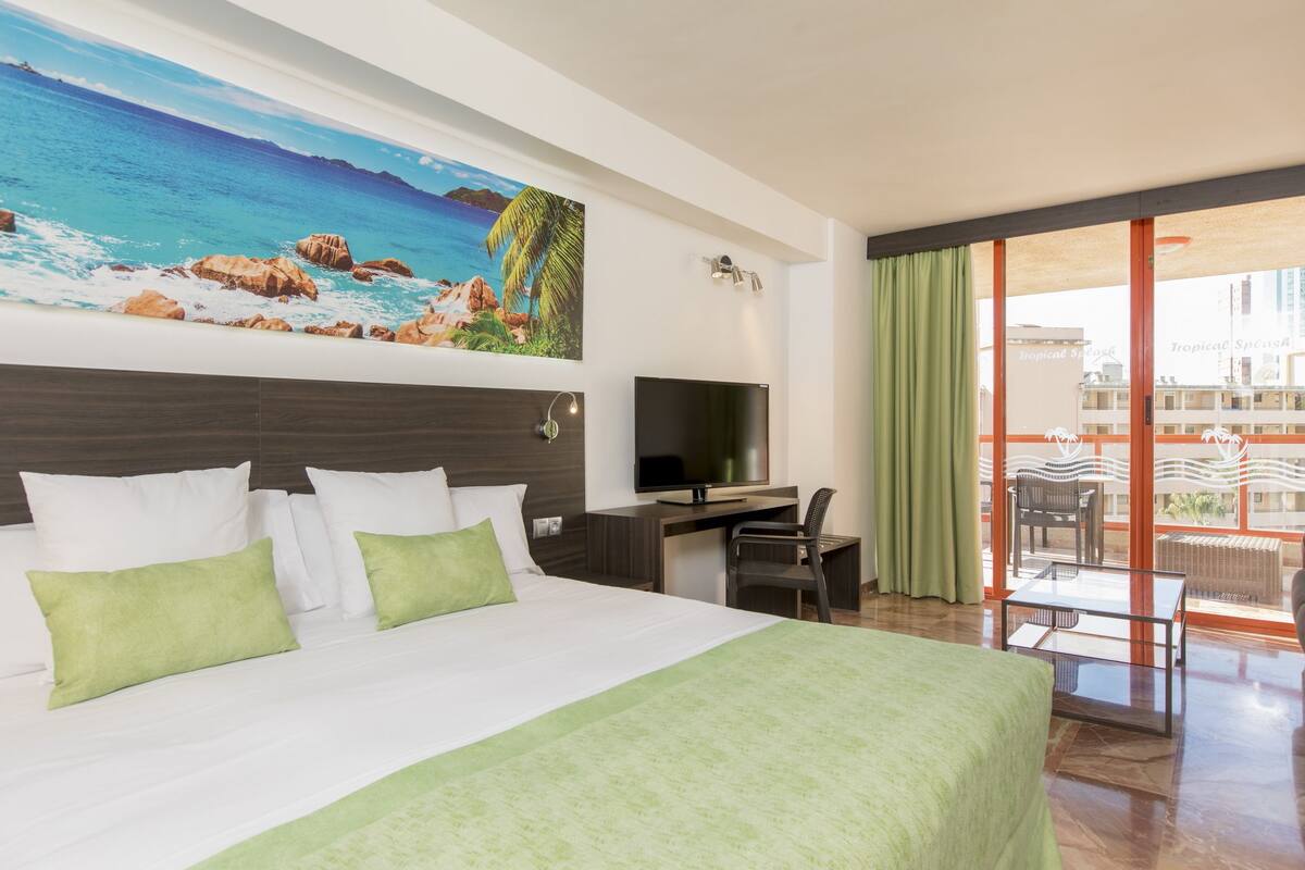 Habitación de hotel con una cama grande, un escritorio con televisor, un balcón con mobiliario exterior y un mural marino en la pared.