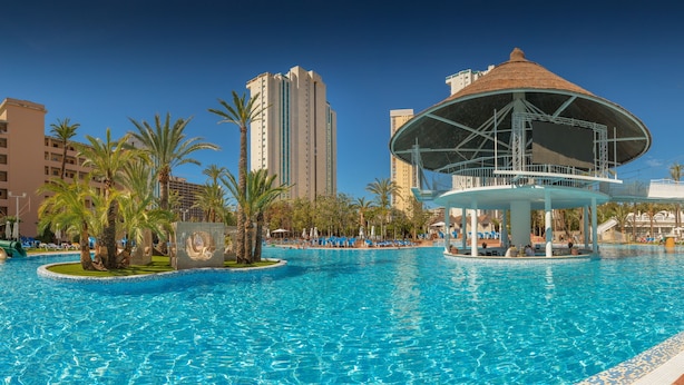 A swimming pool with lounge chairs, palm trees, and a gazebo structure. - Magic Tropical Splash (Finestrat)