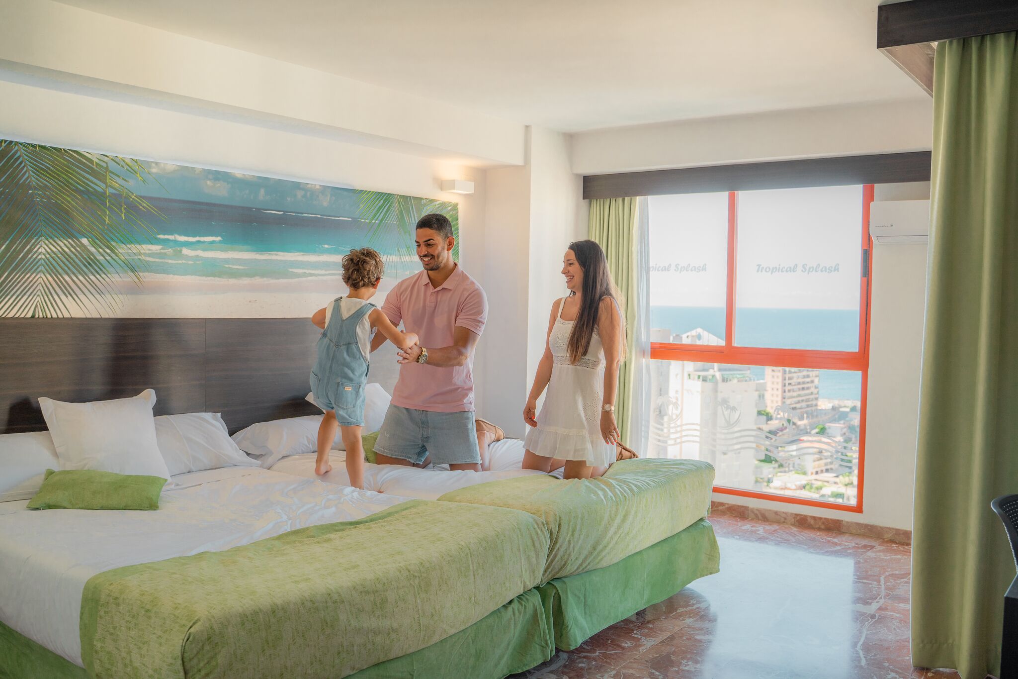 A hotel room with a bed, a large window with a city view, and a mural of a beach scene.