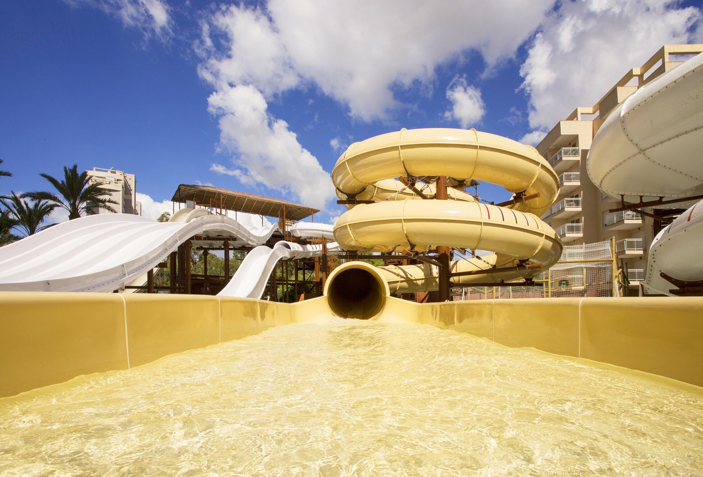 A water slide with a yellow tube leading to a pool, surrounded by buildings and palm trees.