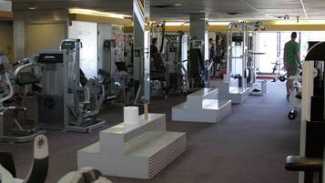 A gym with various exercise machines, a mirrored wall, and a person walking in the background.