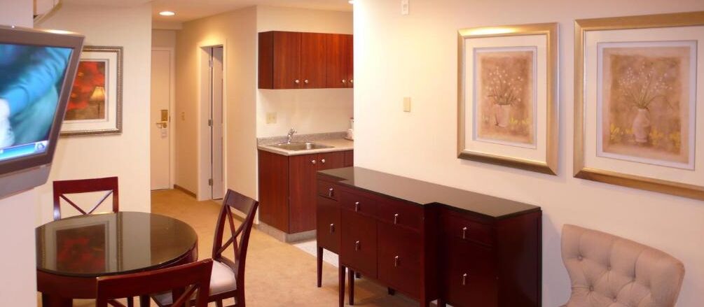A dining area with a round table and chairs, a television, a kitchen with wooden cabinets, and framed artwork on the wall.