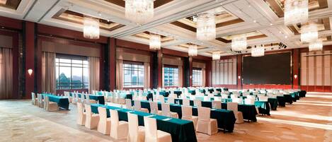 A conference room with rows of tables and chairs, large chandeliers, and a projection screen.