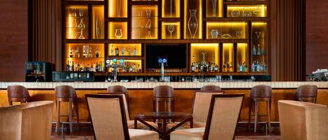 Un'area bar con diversi posti a sedere, una televisione e scaffali con bottiglie e bicchieri di varie forme.