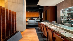 Un área de bar moderna con una barra alargada, sillas de madera y una vitrina de cristal.