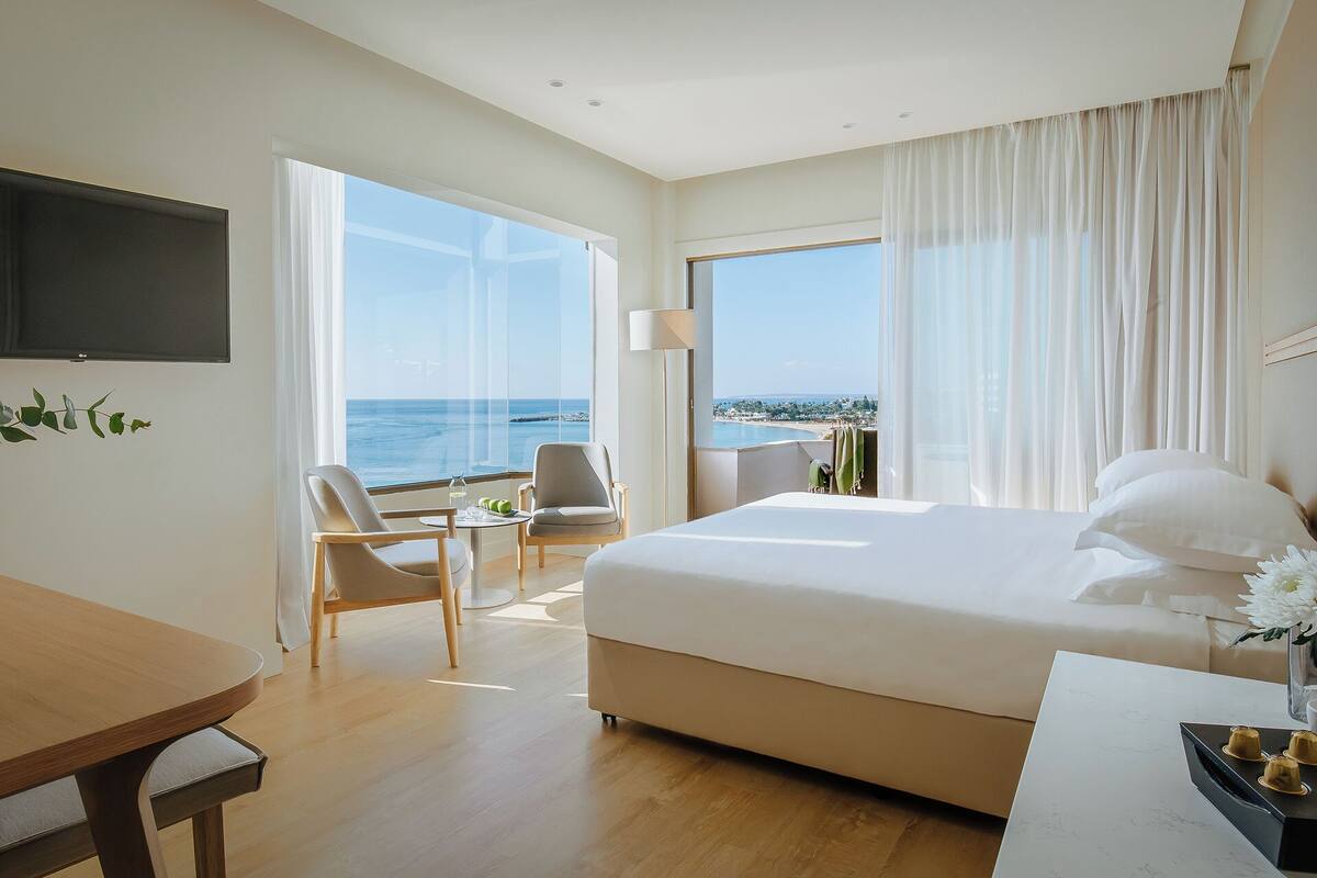 Une chambre d’hôtel avec un grand lit, un bureau, une chaise et une télévision. De grandes fenêtres offrent une vue sur la mer.
