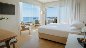 A hotel room with a large bed, a desk, a chair, and a TV. There are large windows with a view of the sea.