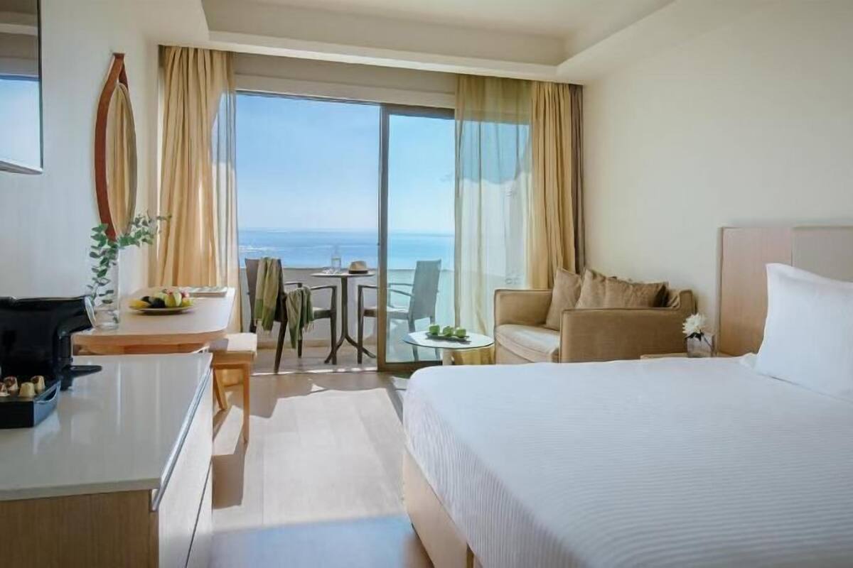 Une chambre d’hôtel avec un lit, un bureau, une chaise, une petite table et une vue sur la mer.