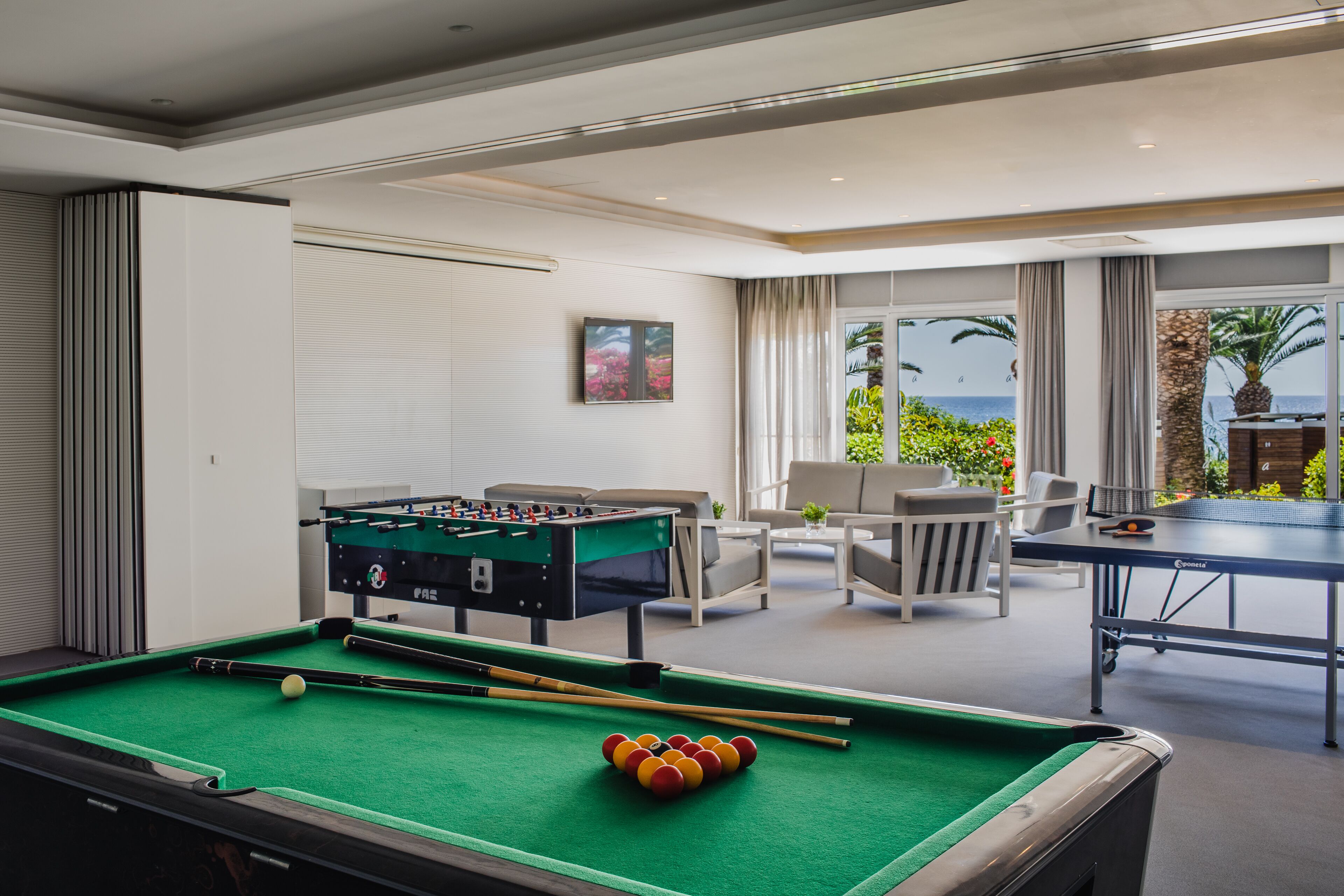 A pool table with a foosball table and a ping pong table in a well-lit room with a view of the ocean.