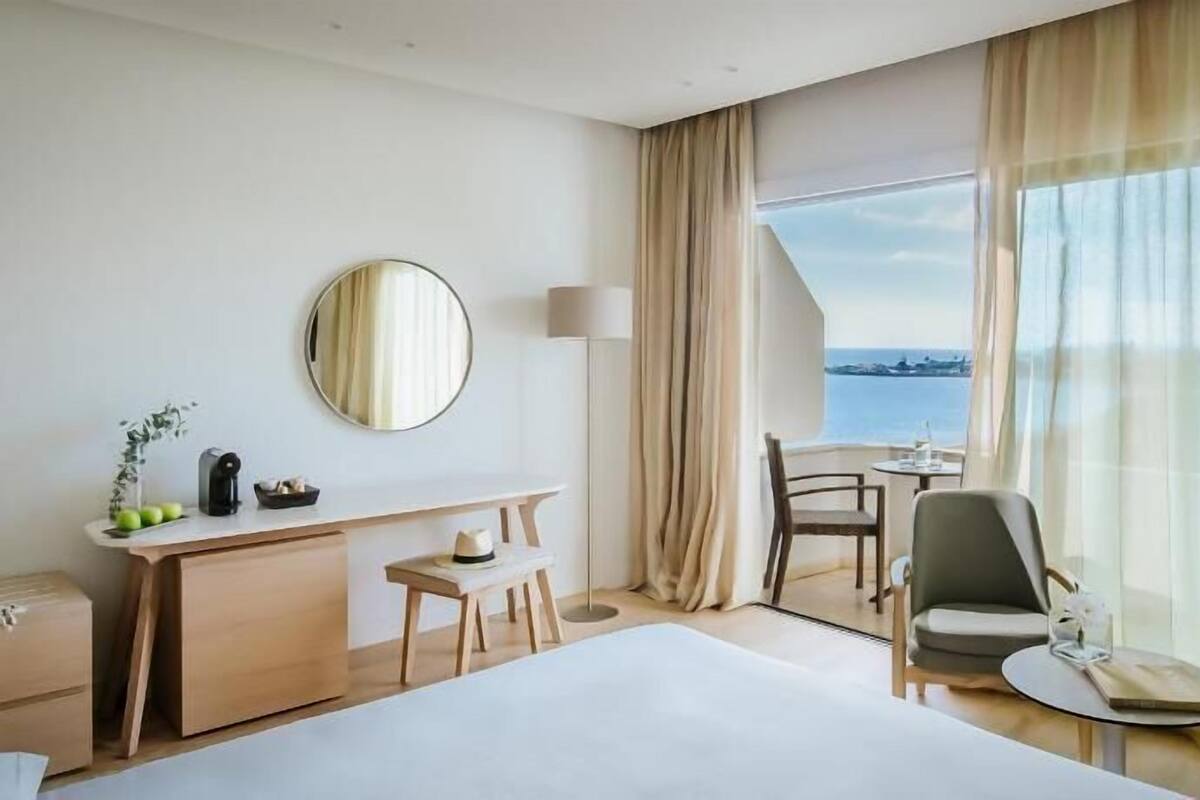Une chambre d’hôtel comprenant un lit, un bureau avec une chaise, une petite table avec une chaise, un miroir rond et une vue sur la mer.