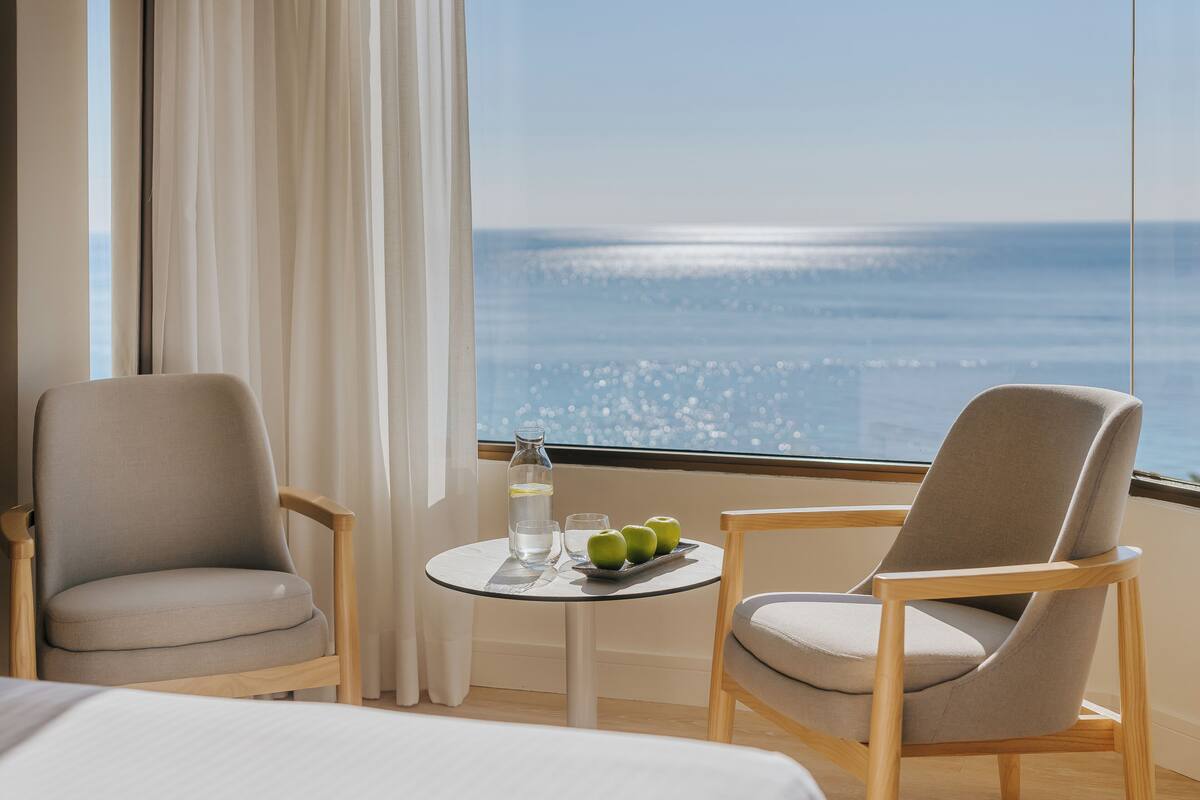 Une pièce avec deux chaises, une petite table sur laquelle se trouvent une bouteille, un verre et des pommes, et une vue sur l’océan.