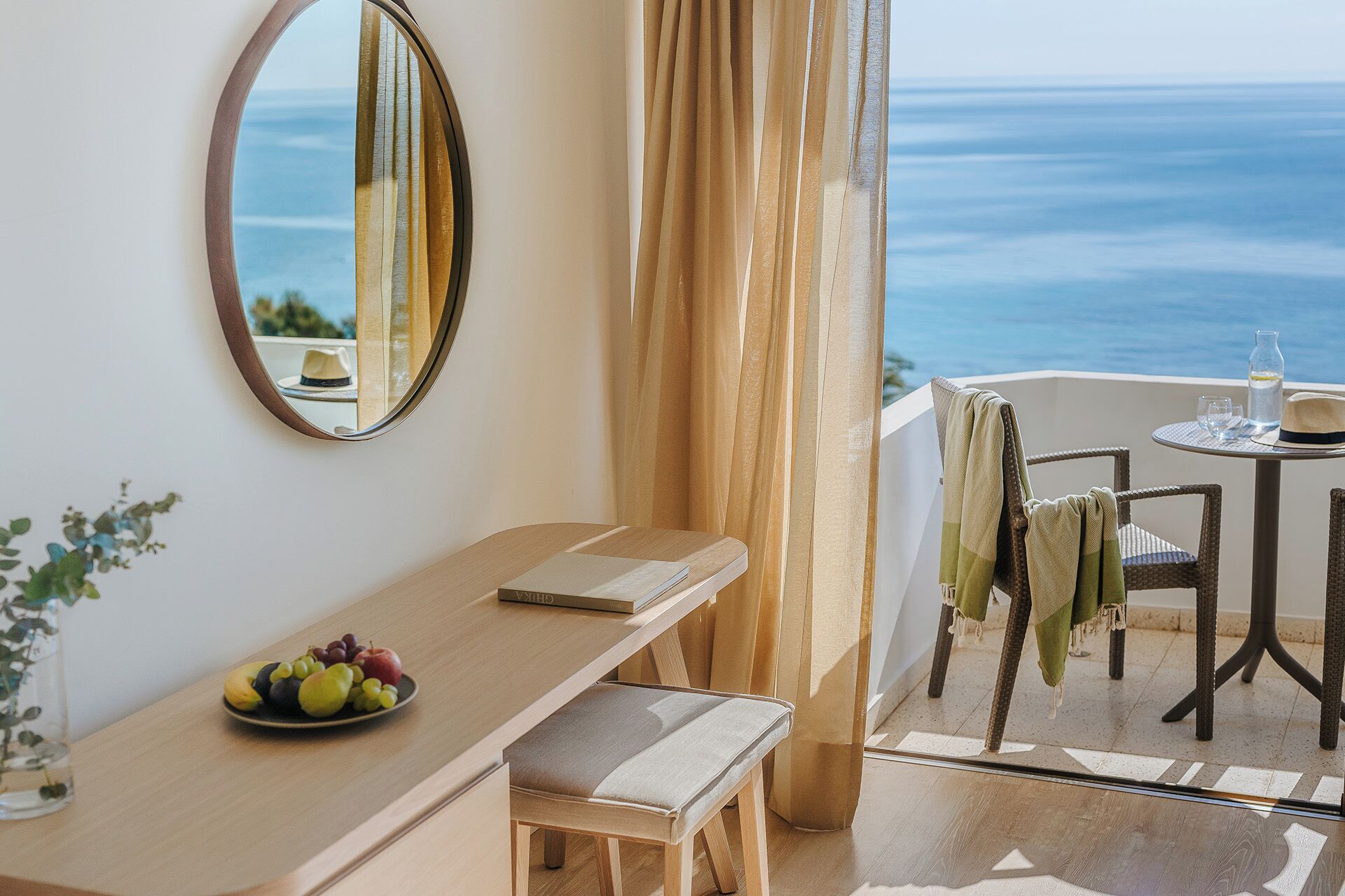 Un balcone con vista sul mare, uno specchio rotondo, un tavolo di legno con un libro e della frutta, e una sedia con un asciugamano.