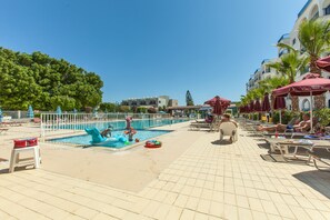 A pool area with lounge chairs and umbrellas, a swimming pool, and a building with balconies in the background. - Crown Resorts Elamaris (Paralimni)