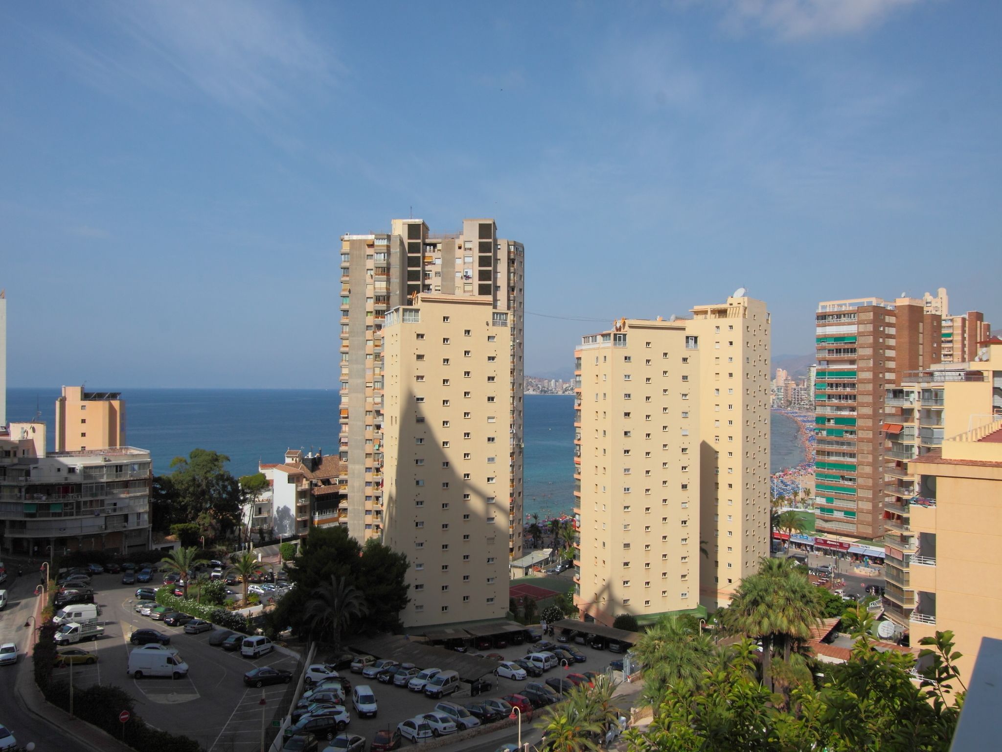 A high-rise building with multiple floors, a parking lot in the foreground, and a view of the sea in the background.