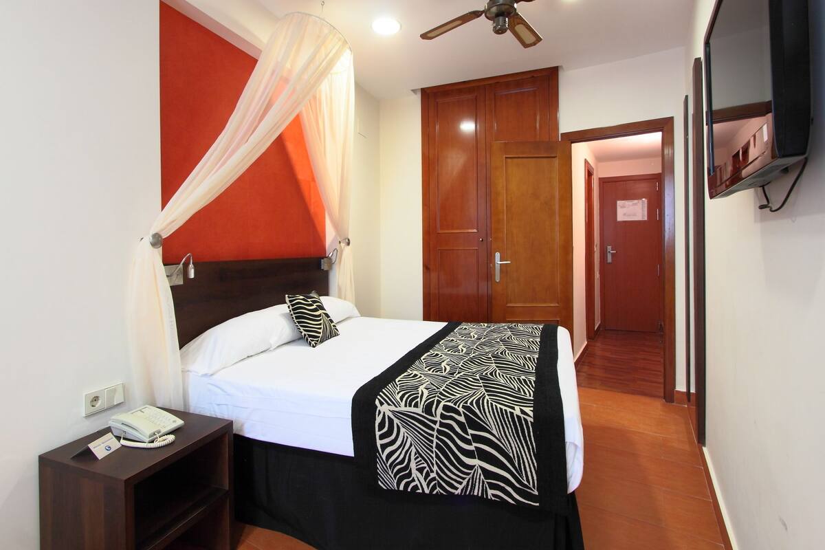 Una habitación de hotel con cama, teléfono junto a la cama, ventilador de techo, televisor montado en la pared y armario de madera.