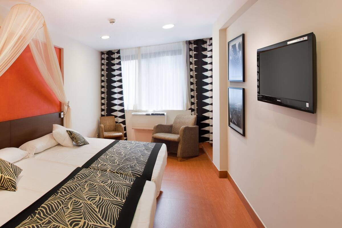 Habitación de hotel con cama, escritorio, silla, televisión y ventana con cortinas.